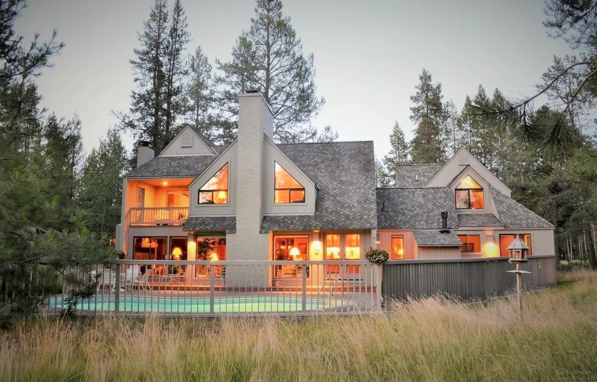 A view of the home's deck with railing, large windows looking out onto the surrounding forest, and abundant greenery all around. The deck invites gathering, relaxation, and easy access to nearby trails or outdoor fun.