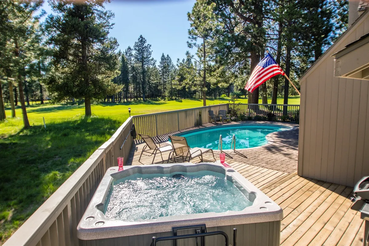 A view of the outdoor pool area and deck, with a hot tub, American flag, and surrounding trees. The scene shows the deck space ideal for summer barbecues and socializing while the kids play in the pool.