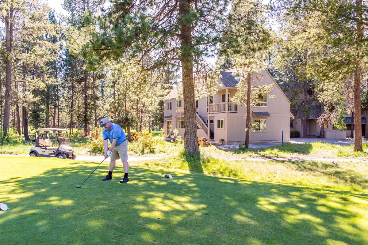 A golfer is preparing to take a shot on a lush green fairway, with a golf cart visible in the background and the house nestled among mature trees. The image captures both the excitement of golf and the residence’s direct relationship to the course.