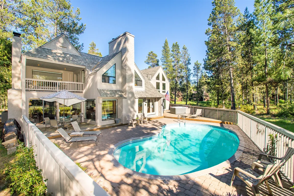 The image features a sparkling private outdoor heated pool framed by comfortable loungers and an inviting outdoor dining area, all set against the beautiful backdrop of a spacious single-family home and lush golf course views. Sunlight glimmers on the water, inviting guests to relax, swim, or gather with friends and family in a serene Sunriver setting.