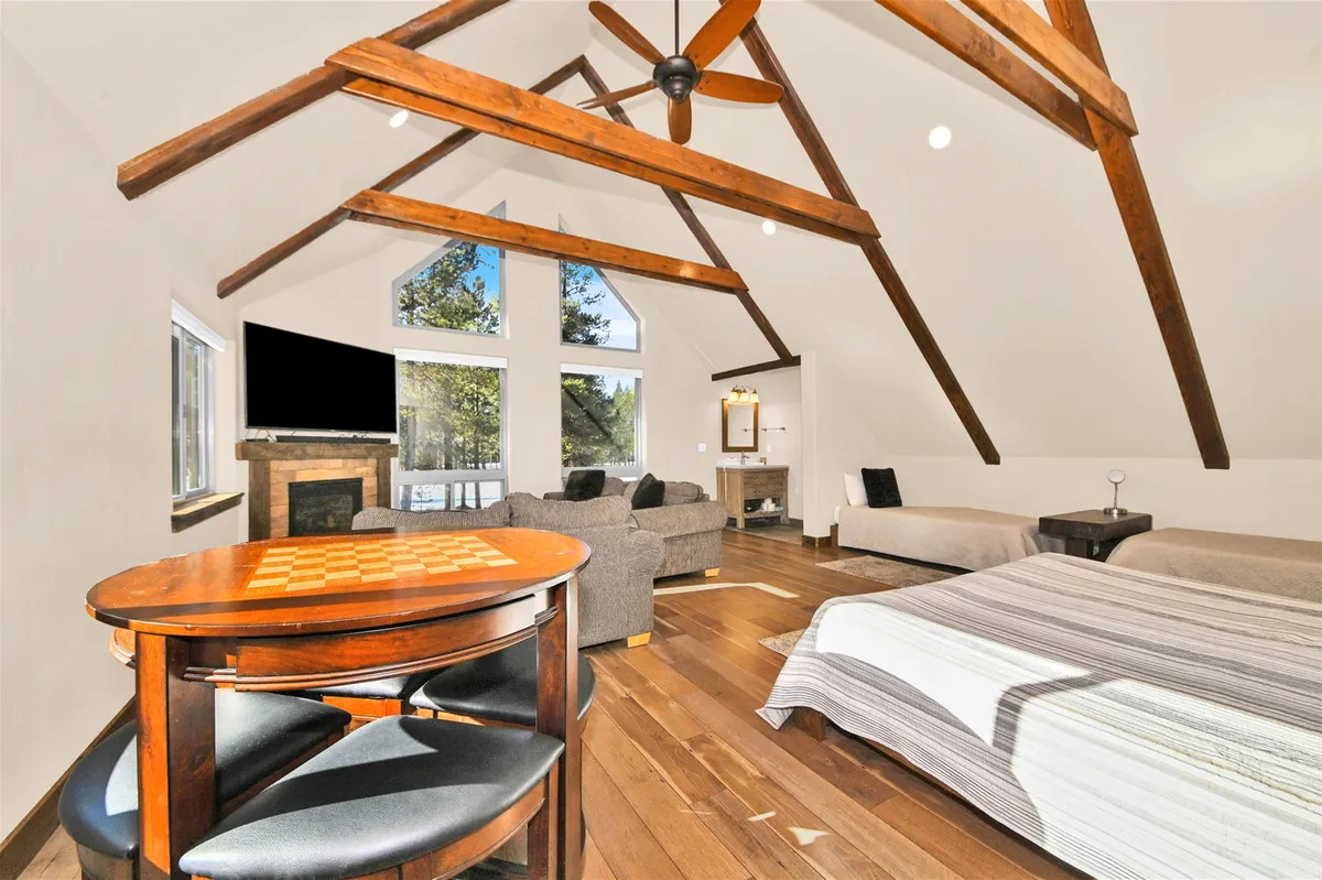 A grand living area with soaring wooden beams, abundant natural light from large windows, and a comfortable seating arrangement. The checkerboard dining table doubles as a space for board games or shared meals. The fireplace and cozy ambiance invite both relaxation and group interaction.