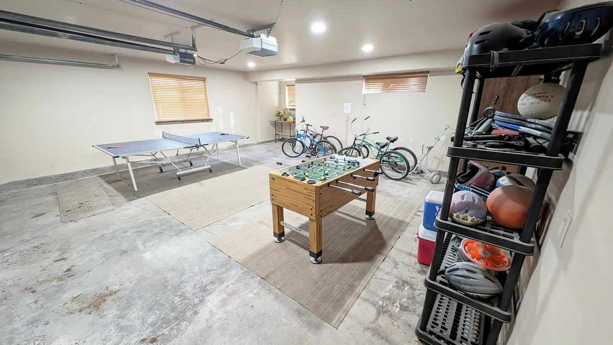 An energetic interior scene featuring a garage game room with a ping pong table, foosball table, bicycles, and sporting equipment. The space is clearly designed for play, recreation, and family fun, all in a relaxed, welcoming atmosphere.