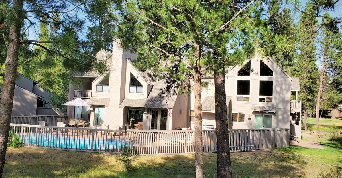 A wide view of the swimming pool and deck area, framed by tall pine trees and the home’s architectural design. The setup invites gatherings, sunbathing, and alfresco dining, with plenty of space for relaxation and social activities.