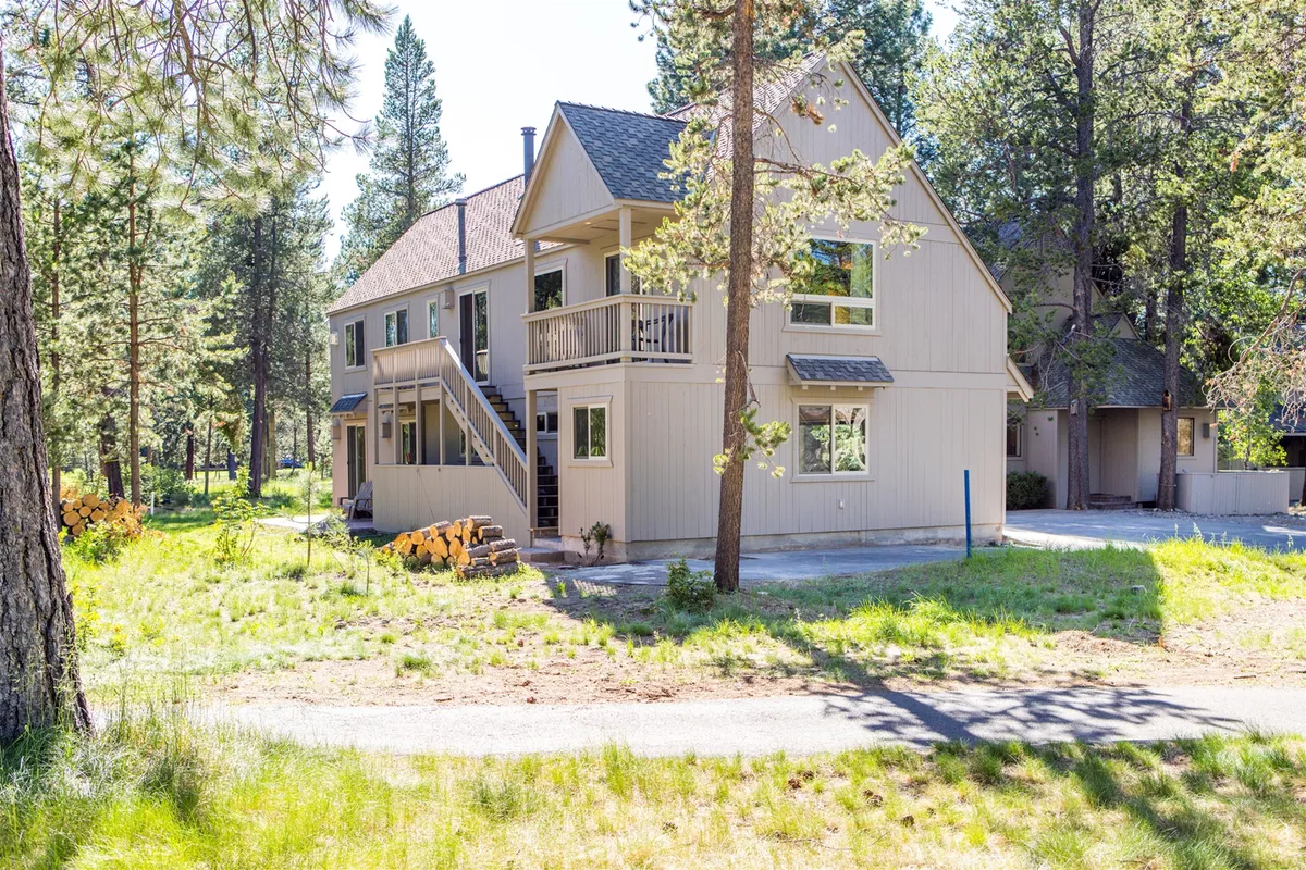 This exterior image shows a two-story house nestled among tall trees, with a welcoming front steps entry, a balcony, and plenty of surrounding greenery. The tranquil setting hints at year-round outdoor enjoyment and the connection to nature that guests can experience.