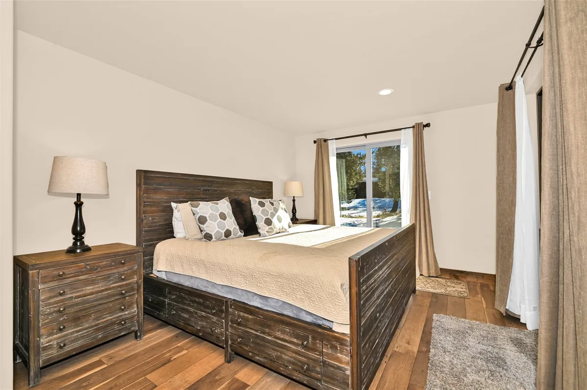 A tranquil bedroom with a wooden bed frame, decorative pillows, and a nightstand with a lamp. A large window provides serene views, inviting natural light and offering a spot for morning coffee or contemplation. The mood is comfortable and private.