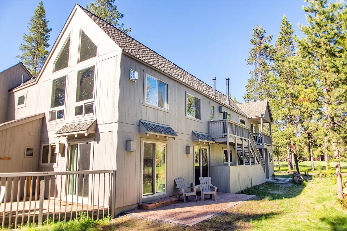 A wooden deck with outdoor seating overlooks a serene landscape of trees, with large windows connecting the indoor and outdoor living spaces. The setting invites relaxation and social gatherings against a backdrop of natural beauty.
